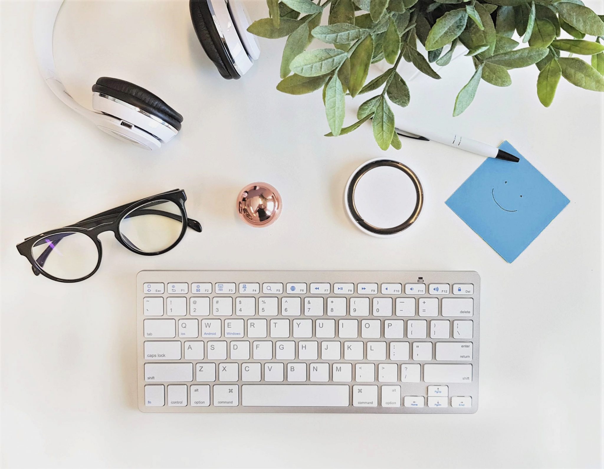 Näppäimistö, silmälasit, kuulokkeet, kasvi ja muistilappu työpöydällä