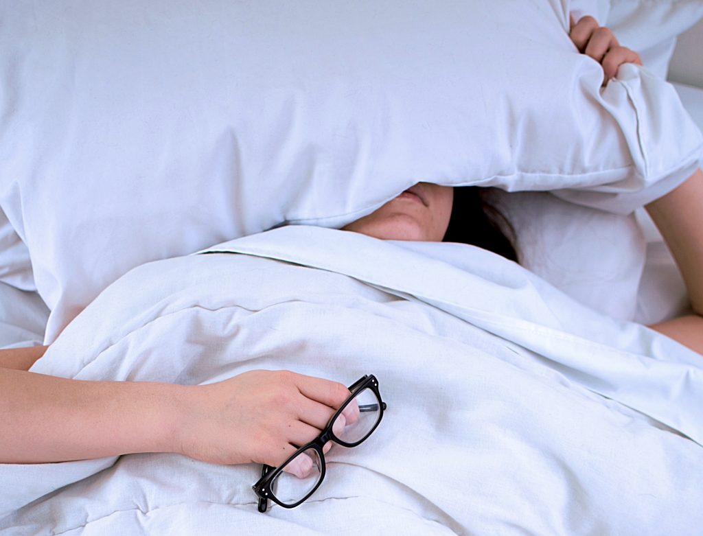 nainen makaa sängyssä väsyneenä pää tyynyn alla ja silmälasit kädessä